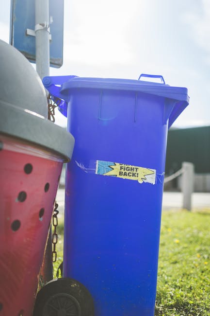 A person's hand is holding a green plastic garbage bag filled with waste or rubbish, with the bag tightly knotted at the top. The bag appears to contain lightweight items, possibly household waste, an