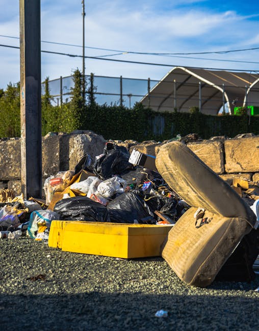 A close-up view of a small shoreline area showing litter and debris collected near a rocky outcrop. The scene features a mix of discarded plastic bottles, paper, and food wrappers scattered among brow